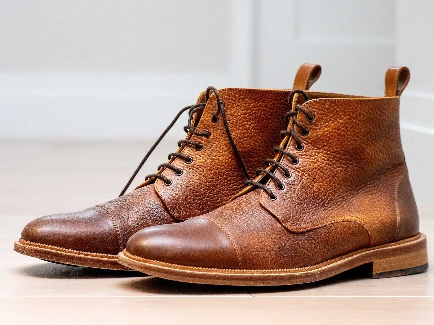 A pair of tan pebble-grain leather lace-up boots with stitched welts on a pale floor