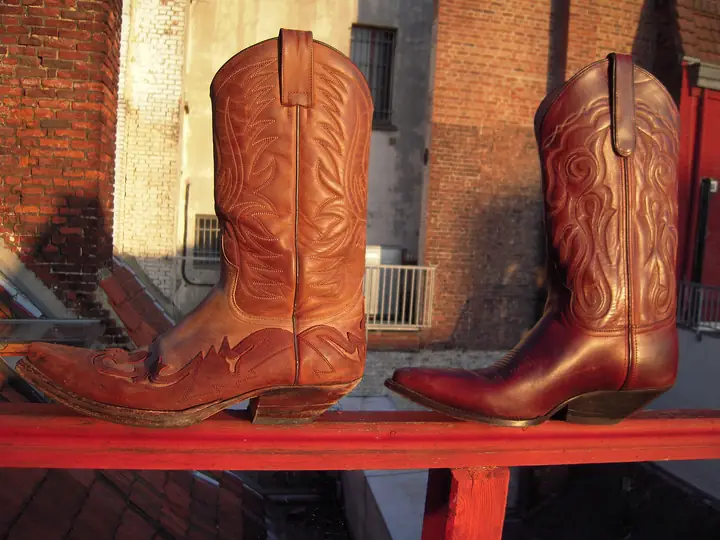 Two tan and brown cowboy boots with stitched shafts standing on a red rail