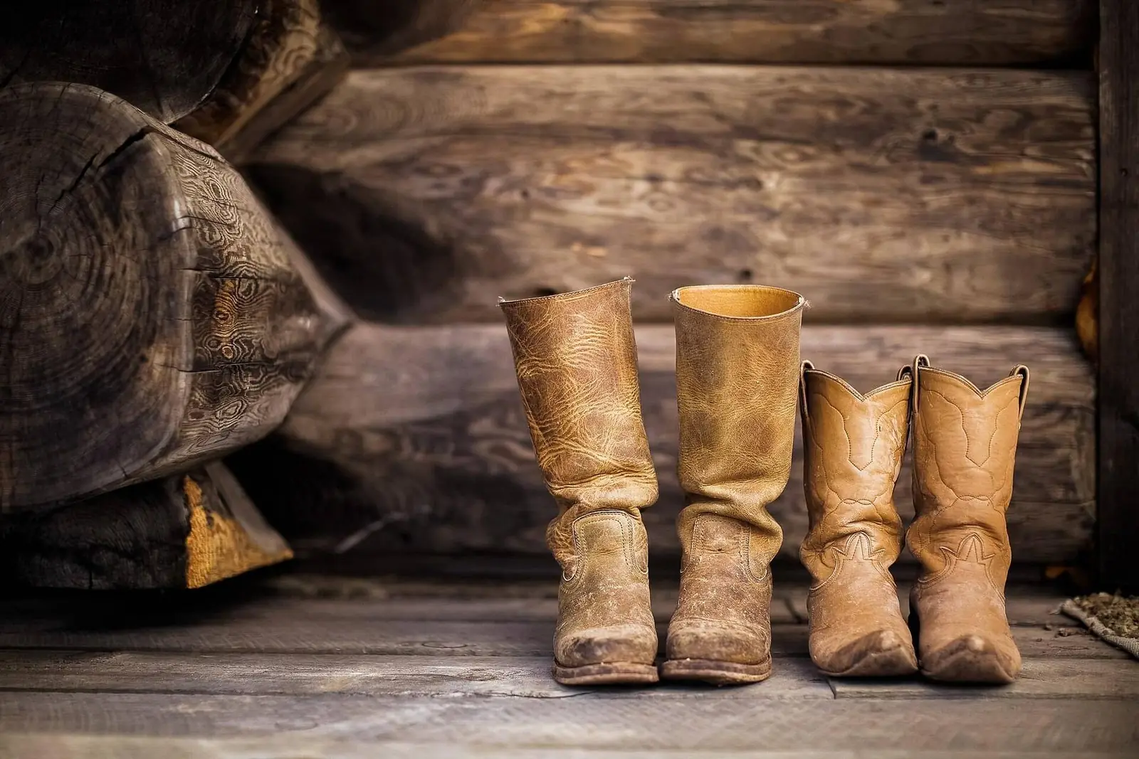 Two worn pairs of tan cowboy boots standing on rough planks against a dark log wall