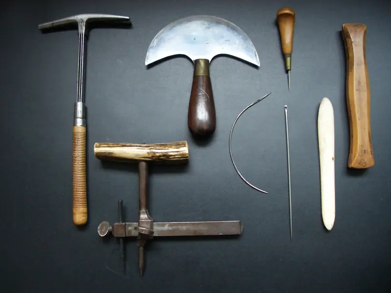 A hammer, a half-moon knife, awls and a curved needle laid out on a dark surface