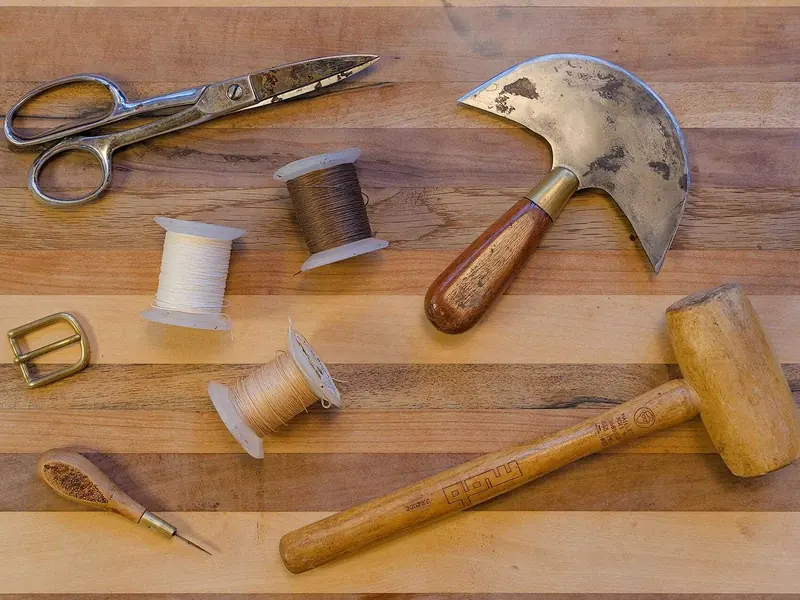 Leatherworking tools on a wooden bench: shears, a round knife, a mallet, thread and an awl