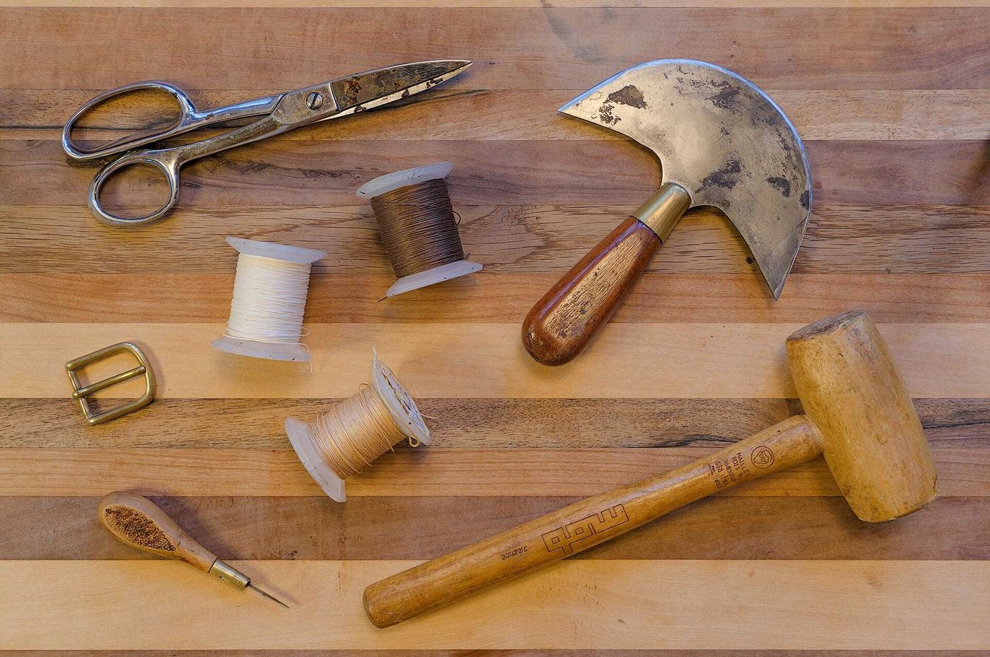 Leatherworking tools on a wooden bench: shears, a round knife, a mallet, thread and an awl
